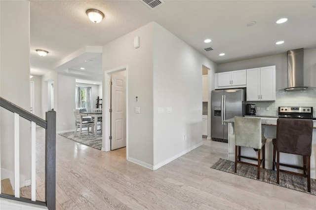 kitchen featuring light hardwood / wood-style floors, wall chimney exhaust hood, a kitchen breakfast bar, white cabinetry, and appliances with stainless steel finishes