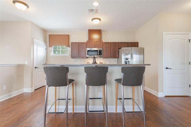 kitchen featuring light stone counters, appliances with stainless steel finishes, dark hardwood / wood-style floors, a kitchen breakfast bar, and a kitchen island with sink