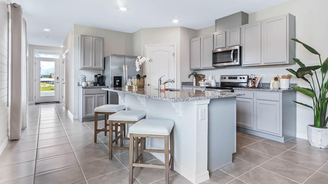 kitchen with stainless steel appliances, gray cabinets, light tile patterned floors, and a kitchen island with sink