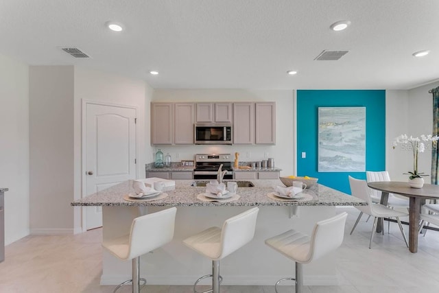 kitchen featuring stainless steel appliances, light stone counters, a textured ceiling, an island with sink, and gray cabinets