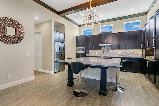 kitchen with beam ceiling, a kitchen island with sink, light hardwood / wood-style flooring, decorative backsplash, and appliances with stainless steel finishes