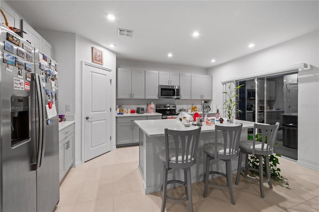 kitchen with appliances with stainless steel finishes, light tile patterned floors, gray cabinets, a kitchen breakfast bar, and a kitchen island with sink