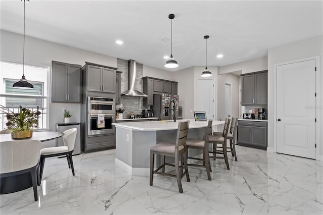 kitchen with stainless steel appliances, wall chimney exhaust hood, hanging light fixtures, an island with sink, and gray cabinetry