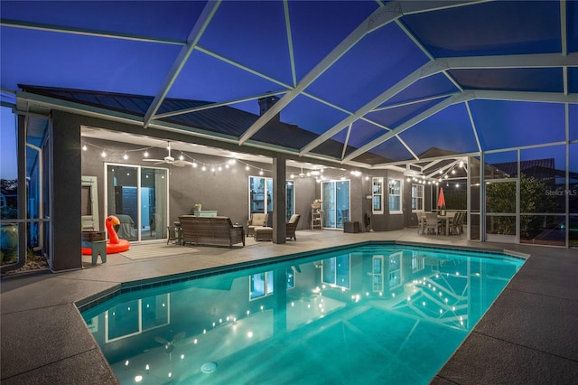 pool at night with a patio, an outdoor hangout area, glass enclosure, and ceiling fan