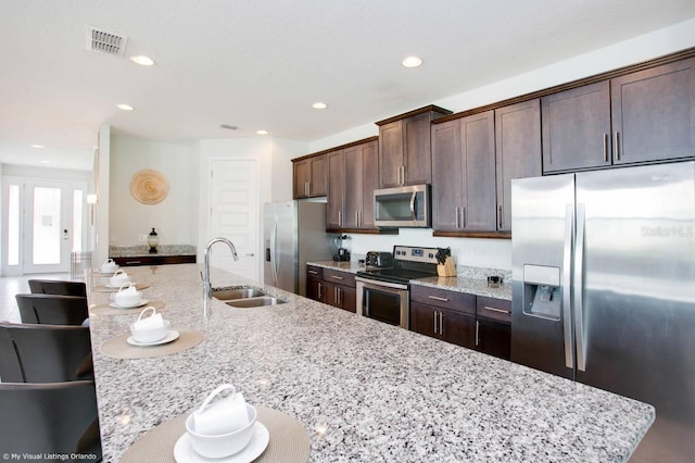 kitchen featuring light stone counters, appliances with stainless steel finishes, dark brown cabinets, sink, and an island with sink