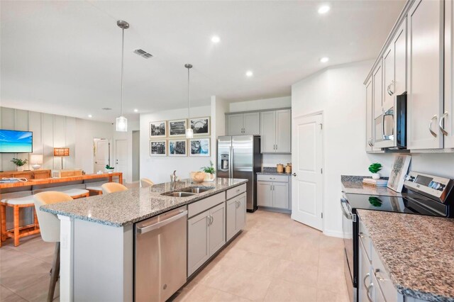 kitchen with sink, light stone counters, appliances with stainless steel finishes, decorative light fixtures, and an island with sink