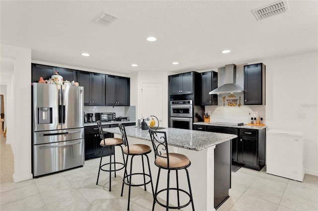 kitchen with a center island with sink, wall chimney exhaust hood, light stone countertops, a kitchen bar, and stainless steel appliances