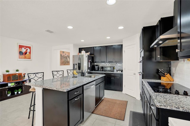 kitchen featuring decorative backsplash, wall chimney exhaust hood, a kitchen island with sink, black appliances, and a breakfast bar area