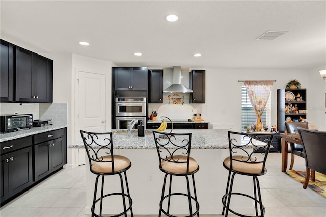 kitchen with light stone counters, a kitchen island with sink, and wall chimney range hood