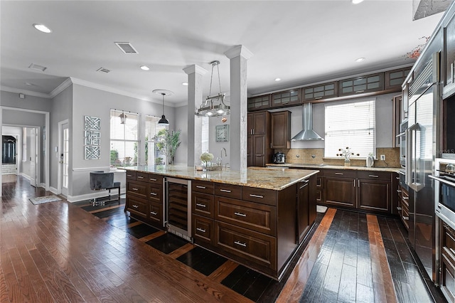 kitchen featuring dark brown cabinetry, hanging light fixtures, beverage cooler, a kitchen island, and wall chimney range hood