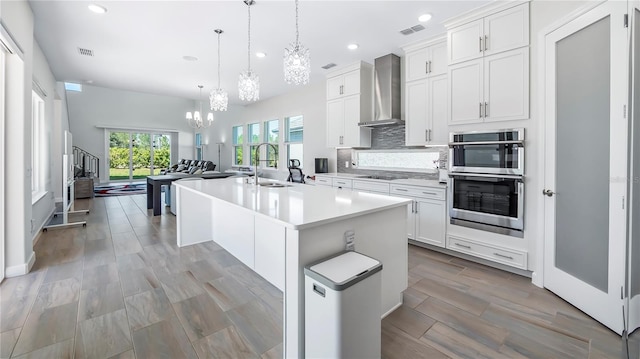 kitchen with pendant lighting, an island with sink, wall chimney range hood, double oven, and white cabinetry