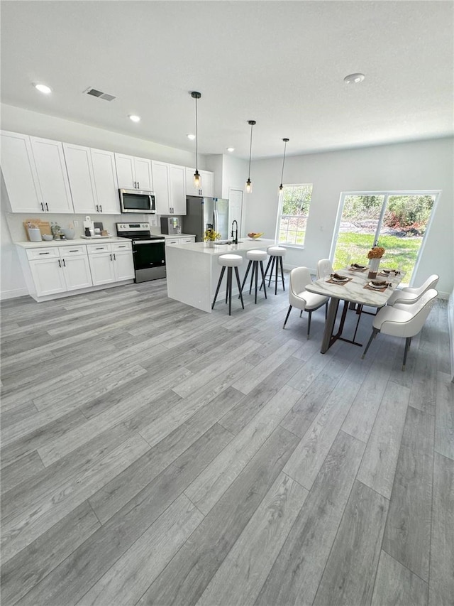 kitchen with white cabinets, light hardwood / wood-style flooring, hanging light fixtures, a kitchen island with sink, and appliances with stainless steel finishes