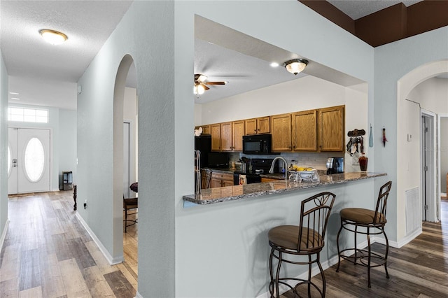 kitchen with dark wood-type flooring, kitchen peninsula, black appliances, a breakfast bar area, and dark stone countertops