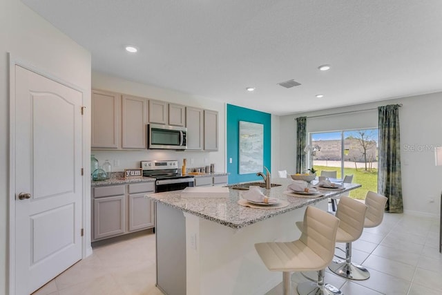 kitchen with sink, light stone counters, appliances with stainless steel finishes, a kitchen bar, and an island with sink