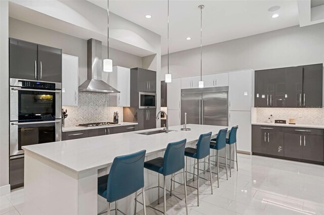 kitchen featuring an island with sink, wall chimney exhaust hood, decorative light fixtures, and built in appliances