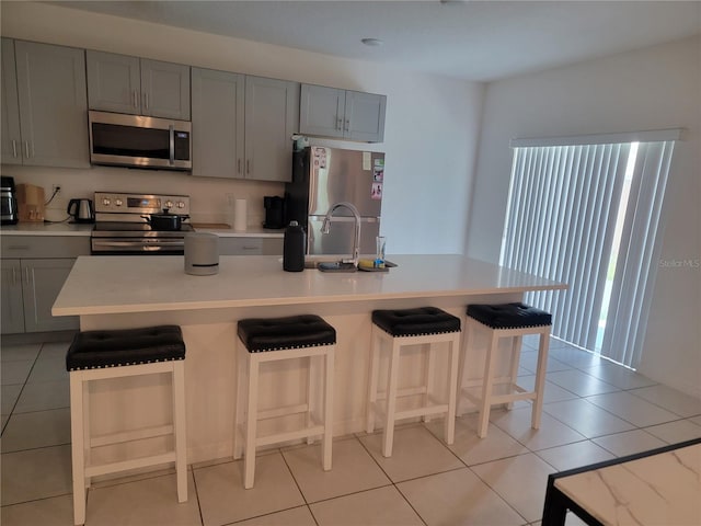 kitchen featuring a center island with sink, stainless steel appliances, light tile patterned floors, gray cabinets, and a breakfast bar area