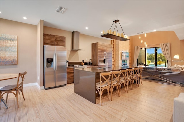 kitchen with stainless steel refrigerator with ice dispenser, wall chimney exhaust hood, hanging light fixtures, a breakfast bar area, and sink