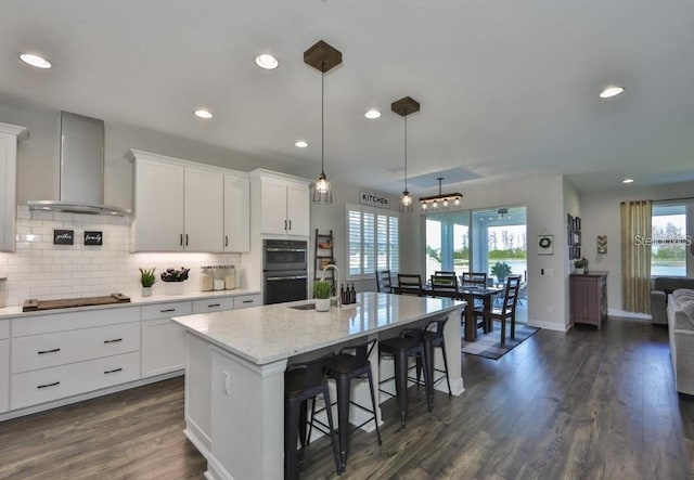 kitchen featuring decorative light fixtures, dark hardwood / wood-style flooring, an island with sink, white cabinets, and wall chimney exhaust hood