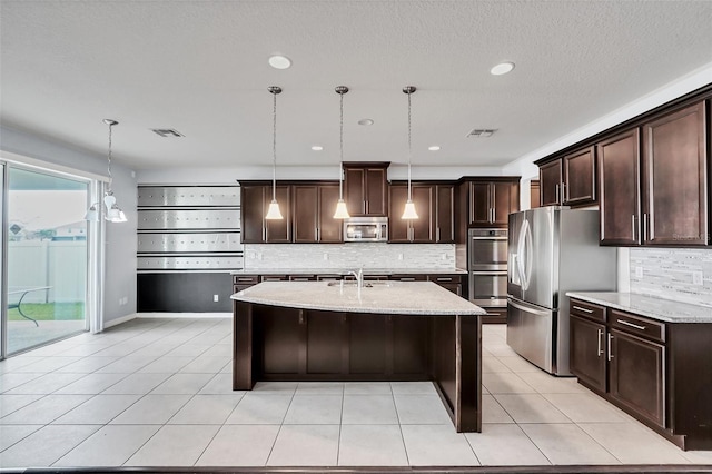 kitchen with appliances with stainless steel finishes, decorative light fixtures, light stone counters, and an island with sink