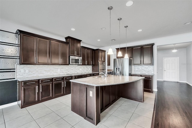 kitchen with pendant lighting, a kitchen island with sink, light hardwood / wood-style flooring, appliances with stainless steel finishes, and dark brown cabinets