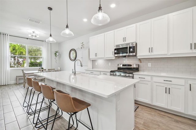 kitchen featuring a center island with sink, appliances with stainless steel finishes, pendant lighting, white cabinets, and light hardwood / wood-style flooring