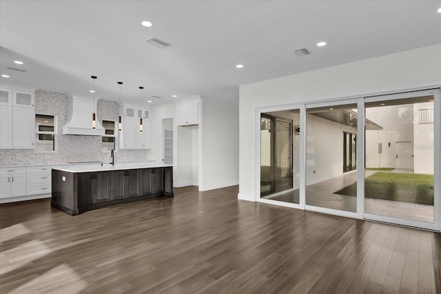 kitchen with custom range hood, white cabinets, a center island with sink, hanging light fixtures, and dark wood-type flooring