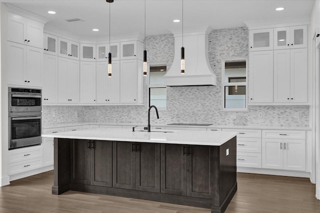 kitchen with a center island with sink, premium range hood, double oven, and dark wood-type flooring