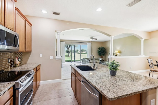 kitchen with a kitchen island with sink, sink, light stone countertops, appliances with stainless steel finishes, and decorative columns