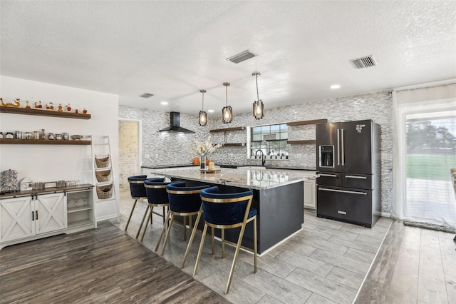 kitchen with high end fridge, wall chimney exhaust hood, a kitchen breakfast bar, light wood-type flooring, and decorative light fixtures