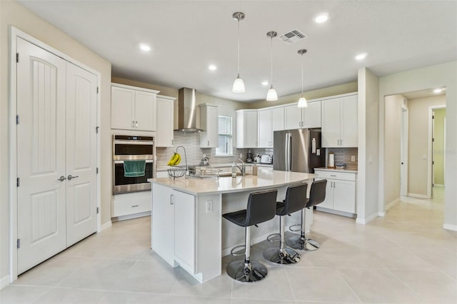 kitchen featuring a center island with sink, white cabinetry, appliances with stainless steel finishes, decorative light fixtures, and wall chimney exhaust hood