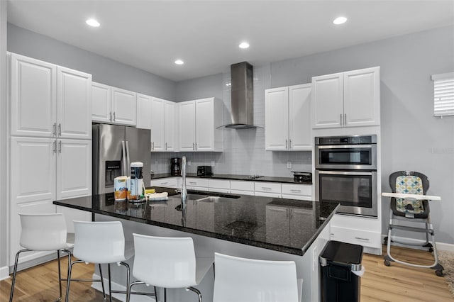 kitchen featuring wall chimney exhaust hood, white cabinetry, an island with sink, and stainless steel appliances