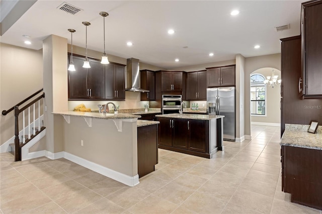 kitchen with kitchen peninsula, appliances with stainless steel finishes, light stone counters, wall chimney exhaust hood, and decorative light fixtures