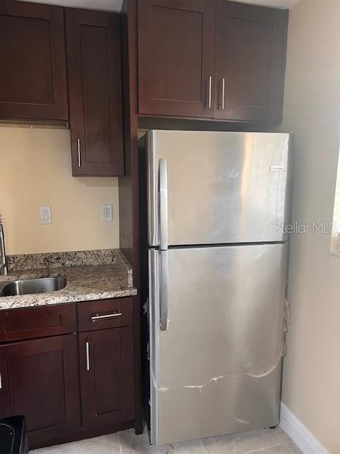 kitchen with light stone counters, dark brown cabinetry, stainless steel refrigerator, light tile patterned floors, and sink