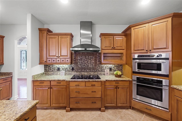 kitchen with light stone countertops, tasteful backsplash, double oven, wall chimney range hood, and light tile patterned flooring