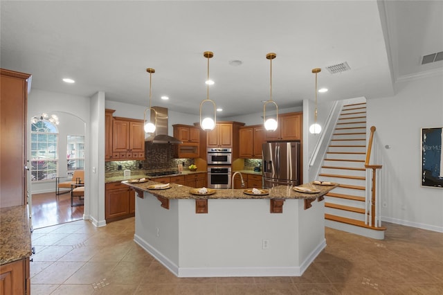 kitchen with appliances with stainless steel finishes, light stone counters, a breakfast bar, wall chimney range hood, and a large island with sink