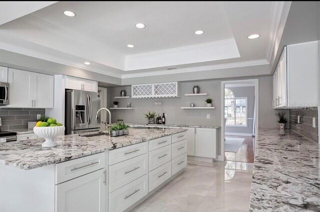 kitchen with a kitchen island with sink, white cabinets, a raised ceiling, appliances with stainless steel finishes, and light stone counters