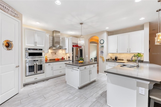 kitchen with white cabinets, wall chimney range hood, sink, hanging light fixtures, and appliances with stainless steel finishes