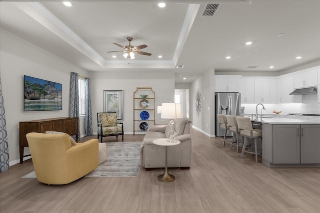 living room featuring sink, crown molding, light hardwood / wood-style flooring, ceiling fan, and a tray ceiling