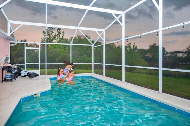 pool at dusk featuring glass enclosure, a patio area, and a lawn