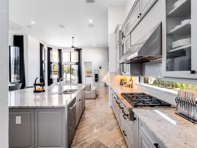 kitchen with sink, an island with sink, wall chimney range hood, light wood-type flooring, and light stone counters