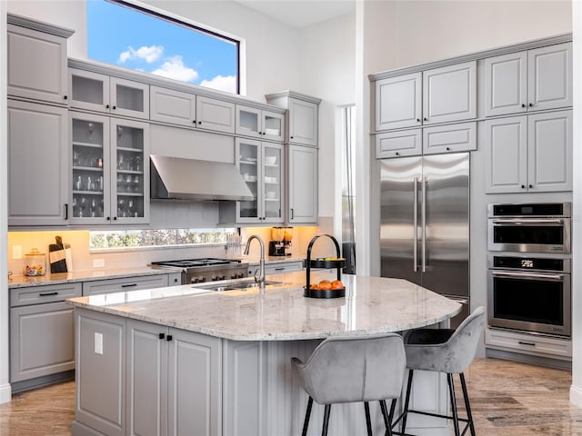 kitchen featuring gray cabinets, an island with sink, wall chimney exhaust hood, and stainless steel appliances