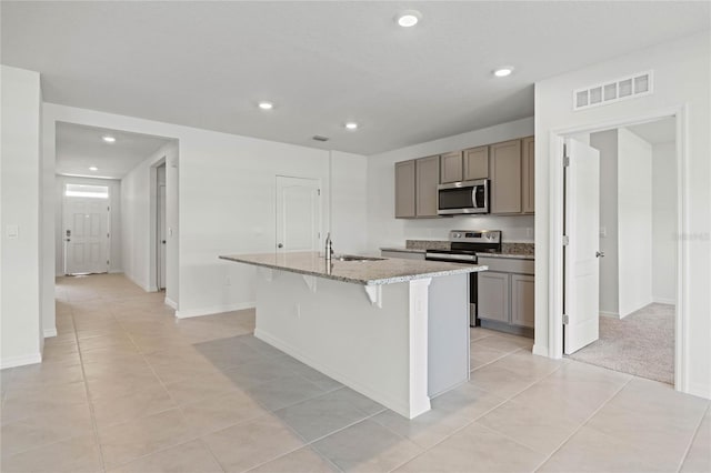 kitchen with stainless steel appliances, a kitchen breakfast bar, light stone counters, an island with sink, and light tile patterned floors