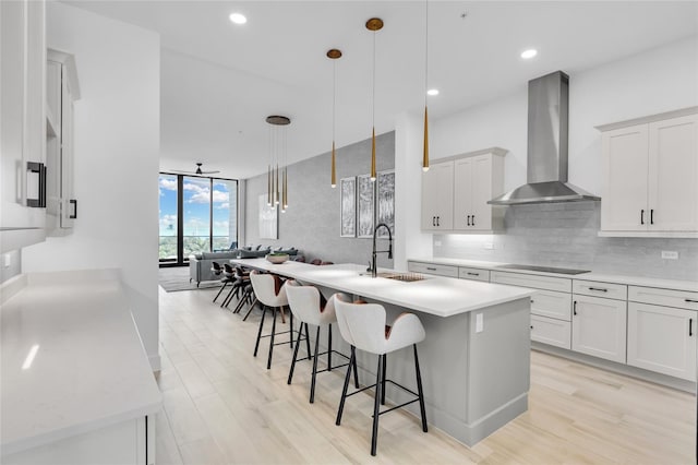 kitchen with white cabinetry, sink, wall chimney exhaust hood, hanging light fixtures, and an island with sink