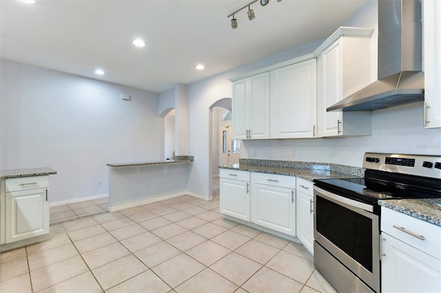 kitchen featuring wall chimney exhaust hood, stainless steel electric range, dark stone countertops, white cabinets, and light tile patterned flooring