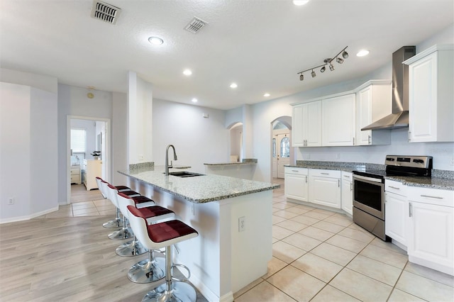 kitchen featuring white cabinets, sink, wall chimney exhaust hood, stainless steel electric range oven, and a kitchen bar