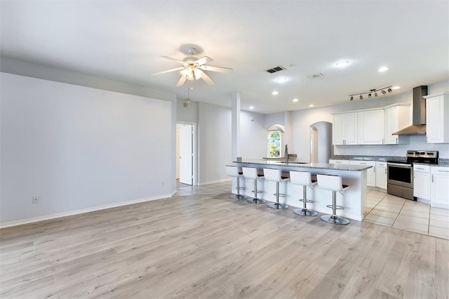 kitchen featuring wall chimney range hood, stainless steel electric stove, a center island with sink, white cabinets, and a kitchen bar