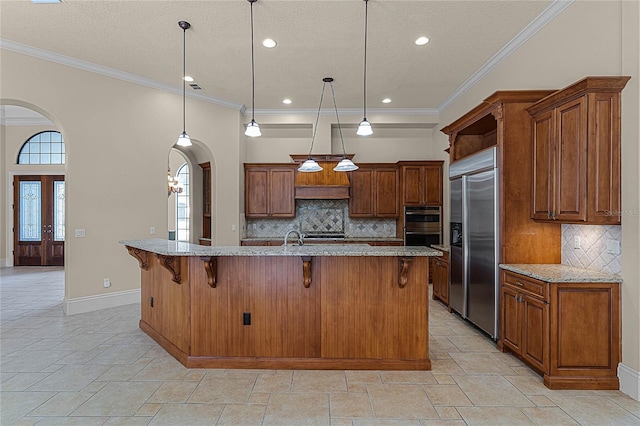 kitchen with stainless steel appliances, hanging light fixtures, light stone counters, a kitchen island with sink, and ornamental molding