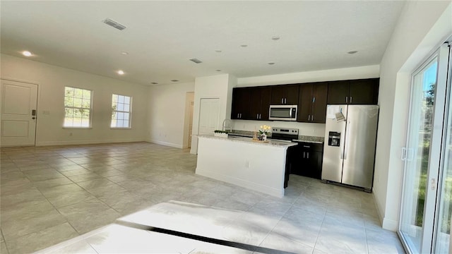 kitchen with dark brown cabinetry, light stone countertops, stainless steel appliances, an island with sink, and light tile patterned flooring