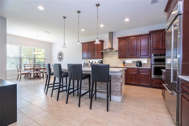 kitchen featuring pendant lighting, wall chimney range hood, an island with sink, appliances with stainless steel finishes, and a breakfast bar area