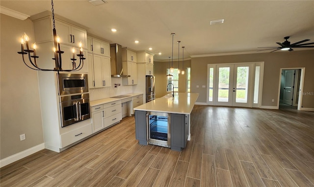 kitchen with a kitchen island with sink, beverage cooler, light countertops, appliances with stainless steel finishes, and wall chimney range hood